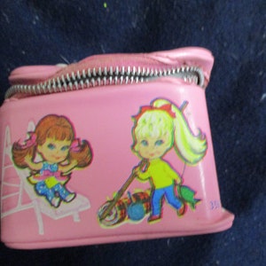 Könnte beinhalten: Rosa Vinyl-Zipper-Tasche mit einem weißen Reißverschluss. Die Tasche zeigt zwei Comic-Mädchen. Ein Mädchen sitzt auf einem weißen Strandstuhl und das andere Mädchen hält eine Harke. Der Text "35" ist auf der Tasche aufgedruckt.