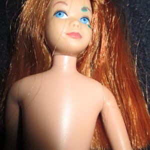 Könnte beinhalten: Eine Vintage-Puppe mit roten Haaren und blauen Augen. Die Puppe hat ein bemaltes Gesicht mit rosa Lippen und einem kleinen grünen Design auf der Wange. Das Haar der Puppe ist lang und seitlich gescheitelt.