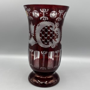 May include: A ruby red glass vase with intricate white etched floral and geometric designs. The vase has a flared top, a rounded base, and a footed bottom. The etched patterns include swirls, dots, and a lattice-like design.