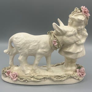 May include: A white ceramic figurine of a girl hugging a donkey. The donkey has a pink flower garland around its neck. The girl is wearing a white dress with pink flowers in her hair. The figurine is set on a base with a straw-like texture and pink flowers.