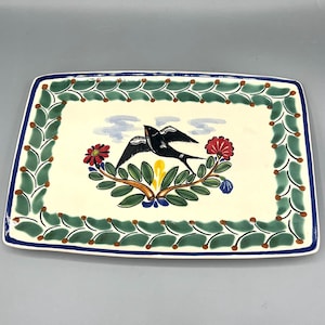 May include: Rectangular ceramic serving platter with a cream-colored center and a green and blue border. The center features a black bird with outstretched wings, red flowers, and green leaves. The border has a repeating leaf design.