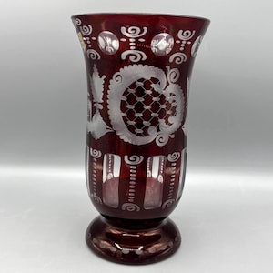 May include: A deep red glass vase featuring intricate white etched patterns, including circular motifs, lattice designs, and vertical panels. The vase has a flared rim and a circular pedestal base, showcasing a contrast between the dark red glass and the frosted white decorative engravings.