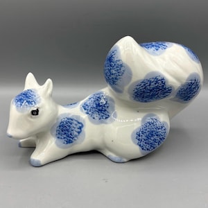 May include: A ceramic squirrel figurine in a reclining pose. The squirrel is white with blue floral patterns on its body and tail. The figurine has black eyes and is a decorative item.