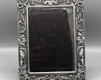 May include: A rectangular picture frame with a dark, textured interior panel. The frame is crafted from a metallic silver-toned material featuring intricate, raised scrollwork, floral motifs, and a decorative border with a fine, linear pattern.