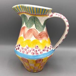 May include: A hand-painted ceramic pitcher with a colorful design. The pitcher features a white base with wavy green, orange, and yellow patterns. The handle is decorated with pink polka dots, and the rim has blue and yellow stripes.