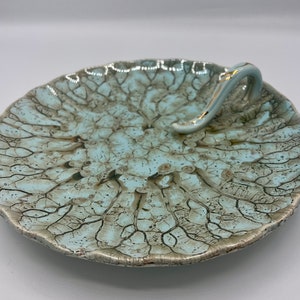 May include: A light blue ceramic serving dish with a crackled glaze and a silver handle. The dish has a unique, organic shape and is perfect for serving snacks or appetizers.
