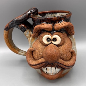 May include: A whimsical ceramic mug with a face design. The mug features a brown handle and a detailed face with a mustache, nose, eyes, and a smiling mouth. The mug is tan and brown with a glossy finish.