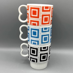 May include: Three stacked white ceramic mugs with geometric square patterns in orange, blue, and black. Each mug has a unique heart-shaped handle. The mugs are stacked on top of each other.