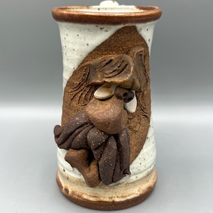 May include: A white and brown ceramic mug with a sculpted face. The face has a brown beard, brown hair, and a brown nose. The mug is glazed with a speckled white glaze.