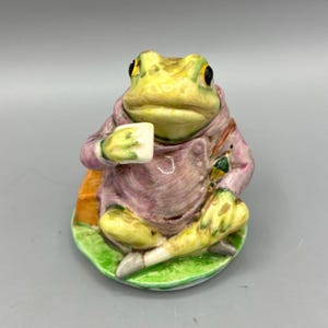 May include: A ceramic frog figurine, dressed in a purple coat, holding a white cup. The frog has yellow and green coloring with black eyes. It is sitting on a green base with a yellow and orange satchel. A whimsical collectible.