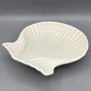May include: A white, seashell-shaped dish with a glossy finish. The dish has a detailed, ridged texture that mimics the natural lines of a seashell. The edges of the dish are gently curved, and the overall design is elegant and coastal-themed.