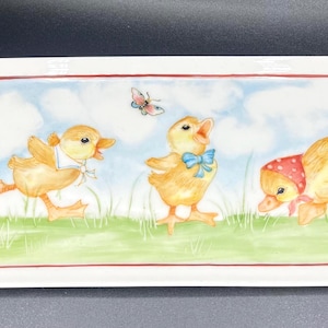 May include: A white rectangular ceramic serving tray with a red border. The tray features a hand-painted design of three yellow ducklings with blue and red accents, a butterfly, and green grass against a blue sky background. The artist's signature is visible in the bottom right corner.