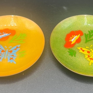 May include: Two oval-shaped decorative dishes. One is yellow with a blue butterfly and red flower design. The other is green with a yellow butterfly and red flower design. Both have a glossy finish.