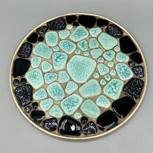 May include: A round decorative plate with a mosaic design. The plate features a gold rim and is filled with turquoise, black, and speckled black ceramic pieces. The turquoise pieces have a crackle glaze.