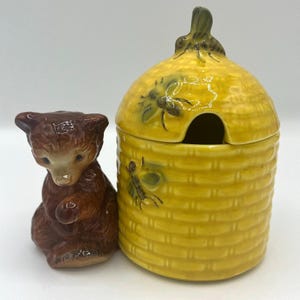 May include: A yellow ceramic honey pot shaped like a beehive with a brown ceramic bear figurine. The honey pot has a lid with a small opening and is decorated with bees.