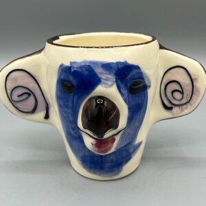 May include: A ceramic mug shaped like a koala's head. The mug is white with blue, brown, and pink painted details. The koala's face is painted with blue fur, a brown nose, and a pink mouth.