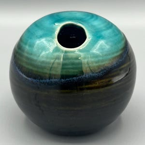 May include: A round ceramic vase with a wide opening. The vase is glazed in shades of teal, green, and black.