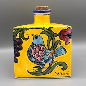 May include: A yellow ceramic bottle with a cork stopper, featuring a hand-painted design of a blue bird, grapes, and flowers. The bottle has a rectangular shape and is signed "Ricceri" on the bottom right.