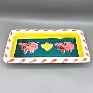 May include: Rectangular ceramic serving dish with a white border adorned with red teardrop shapes. The center features a teal background with two pink pigs and a yellow heart. A yellow border frames the teal section.