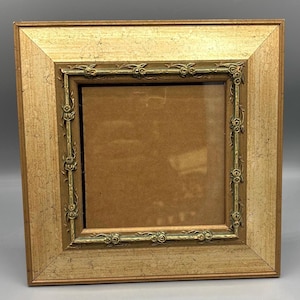 May include: Square gold-toned picture frame with a decorative inner border. The frame has a textured finish and a square opening for a photograph or artwork. The inner border features a floral design, adding a touch of elegance to the frame.