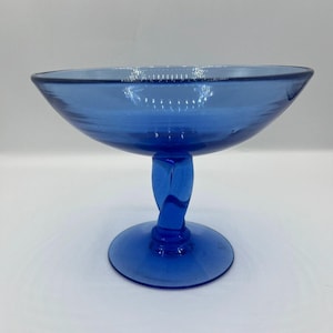 May include: A cobalt blue glass compote dish featuring a wide, shallow bowl with horizontal ridges along the interior and a central pedestal base with a distinct twisted stem design, supported by a circular foot for stability and balance.
