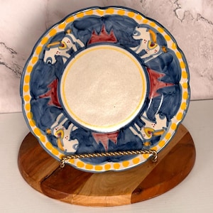 May include: A round ceramic plate with a scalloped edge featuring a central cream-colored circle surrounded by a decorative border of blue and red painted patterns, finished with a yellow dotted rim against a white background.