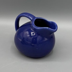 May include: A cobalt blue ceramic pitcher with a rounded body and a curved handle. The pitcher has a spout for pouring and is likely used for serving liquids. The glossy finish reflects light.