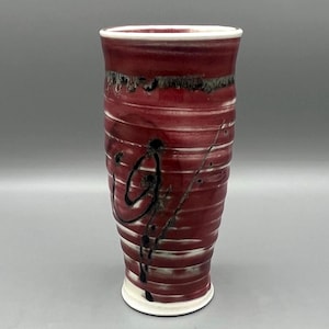 May include: A red ceramic vase with black and white abstract designs. The vase has a cylindrical shape and a flared rim.