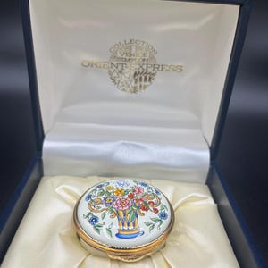 May include: A white porcelain trinket box with a gold trim and a floral design. The box is in a blue velvet box with the words "Collection Venice Simplon Orient Express" printed on the inside.