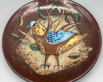 May include: A brown ceramic plate with a stylized painting of a bird in a tree. The bird is blue, yellow, and green, and the tree is brown with green leaves. The plate has a rustic, hand-painted look.