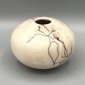 May include: A small, round, off-white ceramic vase with a narrow opening. The vase features dark, linear crackle patterns across its surface, creating a unique, textured appearance. The vase is a decorative item.