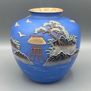May include: A blue ceramic vase with a raised design of a Japanese landscape. The vase features a pagoda, a tree, mountains, and birds in flight. The vase has a light blue glaze with accents of gold, red, and green.