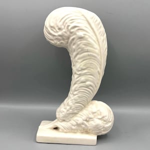 May include: A cream-colored ceramic feather sculpture. The feather is detailed with textured ridges and curves, mounted on a rectangular base. The sculpture has a matte finish and is set against a gray background.