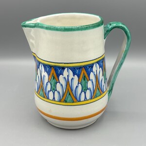 May include: A white ceramic pitcher with a green handle and rim. The pitcher features a decorative band of blue, yellow, and green floral patterns. An orange band encircles the base.