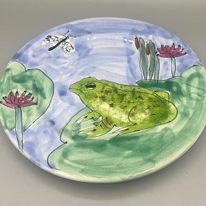 May include: A hand-painted ceramic plate featuring a green frog sitting on a lily pad with a dragonfly flying above. The plate has a blue background with green lily pads and pink flowers.