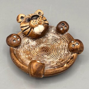 May include: A round ceramic trinket dish featuring a sculpted tiger head at the top and three tiger paws extending from the sides. The dish has a textured, spiral-patterned interior in shades of brown and tan with a glossy finish.