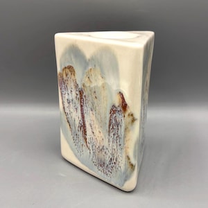 May include: A triangular ceramic vase with a smooth, off-white base glaze. The surface features a reactive drip pattern in shades of muted blue, charcoal gray, and deep reddish-brown, creating a textured, layered appearance across the three sides.