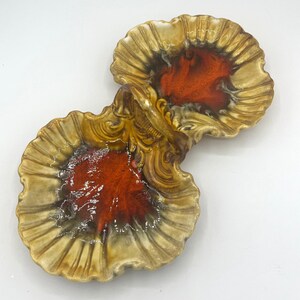 May include: A ceramic serving dish with two sections, each shaped like a scallop shell. The dish is glazed in shades of brown, orange, and white, with a decorative handle in the center.