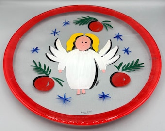 May include: A clear glass plate with a red rim, featuring a painted angel with yellow hair, white wings, and a white robe. The plate is decorated with red berries, green sprigs, and blue stars. The text "KOSTA BODA" is visible.