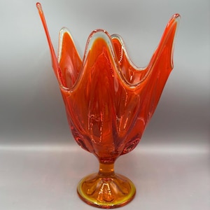 May include: A vibrant orange glass vase with a unique, organic shape. The vase features a flared, petal-like rim and a pedestal base. The glass has a glossy finish, with a clear rim and a yellow base.