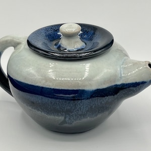 May include: A white and blue ceramic teapot with a lid. The teapot has a rounded body and a spout on the side. The lid is a separate piece and has a blue glaze.
