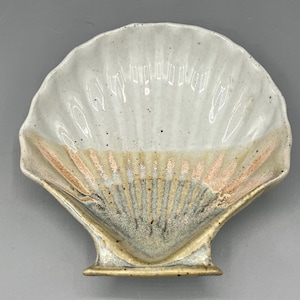 May include: A white ceramic seashell dish with a light brown and pink glaze. The dish is shaped like a scallop shell and has a raised base.