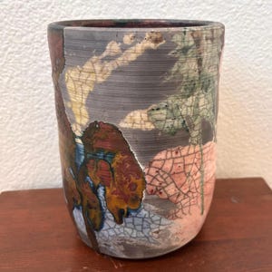 May include: A cylindrical ceramic cup featuring a raku-style finish with a prominent crackle glaze pattern. The surface displays a mix of dark charcoal, muted green, earthy brown, and soft pink tones, with horizontal striations throughout.