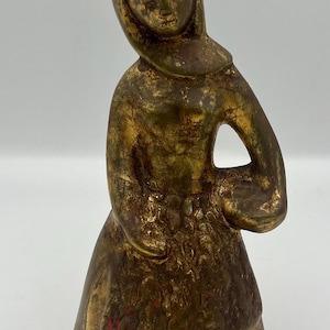 May include: A gold-painted ceramic figurine of a woman wearing a hooded dress and holding a basket. The figurine has a rustic, aged appearance.