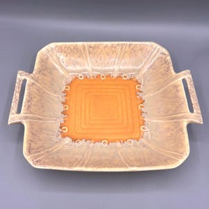 May include: A square, vintage serving dish with a tan and orange color scheme. The dish has a raised, textured design with a central orange square and a series of concentric squares. It features integrated handles on either side.