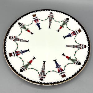 May include: A white ceramic plate with a gold rim and a black scalloped edge. The plate features a repeating pattern of colorful nutcracker figures with a garland of green leaves and red berries. The nutcrackers are in various colors, including red, blue, and green.