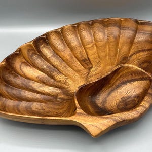 May include: A wooden serving dish with a shell-like design. The dish is made of a light brown wood with darker brown grain patterns. It has a large, fan-shaped section and a smaller, oval-shaped section, perfect for serving snacks or appetizers.