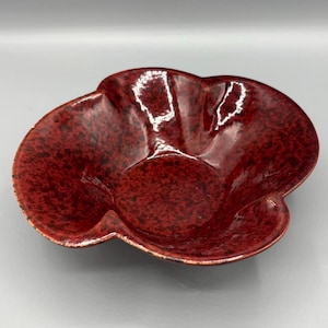 May include: A red speckled ceramic bowl with a unique, organic shape. The bowl has a glossy finish and a dark red color with black speckles. The rim of the bowl has a lighter, more faded red color.