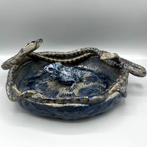 May include: A ceramic bowl with a blue and gray lizard sculpture inside. The bowl has a textured surface and is surrounded by a snake-like rim. The lizard is detailed with blue and white accents.