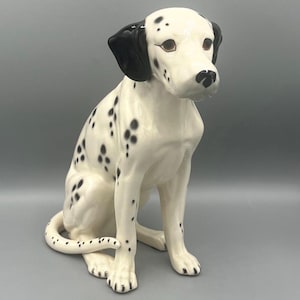 May include: A ceramic Dalmatian dog figurine. The dog is sitting with its tail curled. It has a white body with black spots, black ears, and brown eyes. The figurine is a decorative item.